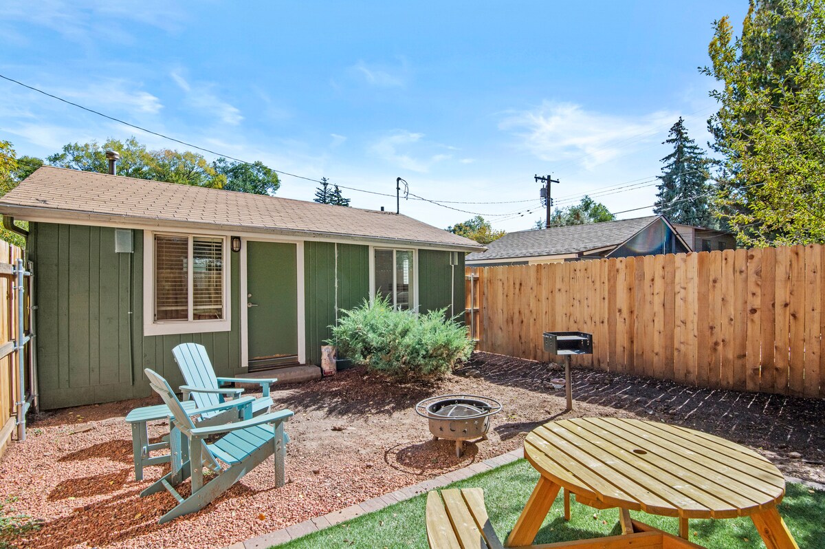 A fully fenced backyard features a picnic table and a fire pit, surrounded by gravel and grass. Two Adirondack chairs are placed nearby, offering a relaxing space. The bungalow is visible in the background, with green walls and a door leading inside.