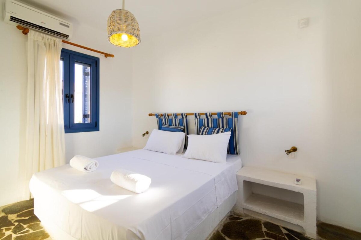 A bright bedroom features a spacious bed with crisp white linens and two rolled towels. Blue and white striped headboards add a touch of color. Natural light fills the room through a window dressed with light curtains, and a woven pendant lamp hangs overhead.