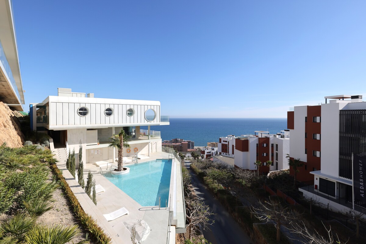 The image features a modern apartment building with a large swimming pool in the foreground, overlooking the ocean. Lush greenery and neatly arranged landscaping surround the pool area, while the building's design includes distinctive circular windows and clean lines, creating a contemporary aesthetic.