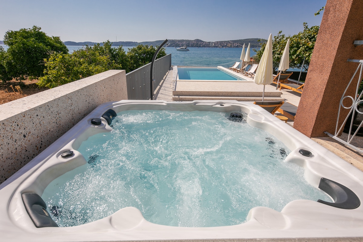 A hot tub is positioned on a terrace with a view of the Adriatic Sea. The bubbling water is surrounded by plants and a swimming pool is visible in the distance. Sun loungers and umbrellas are arranged neatly near the pool, enhancing the outdoor relaxation space.