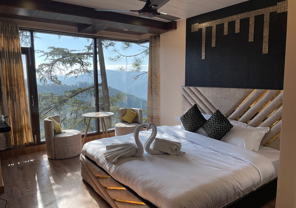 A well-appointed room features a spacious bed with decorative towels arranged in the shape of swans. A small round table is positioned next to two stylish chairs, with large windows offering a mountain view in the background.