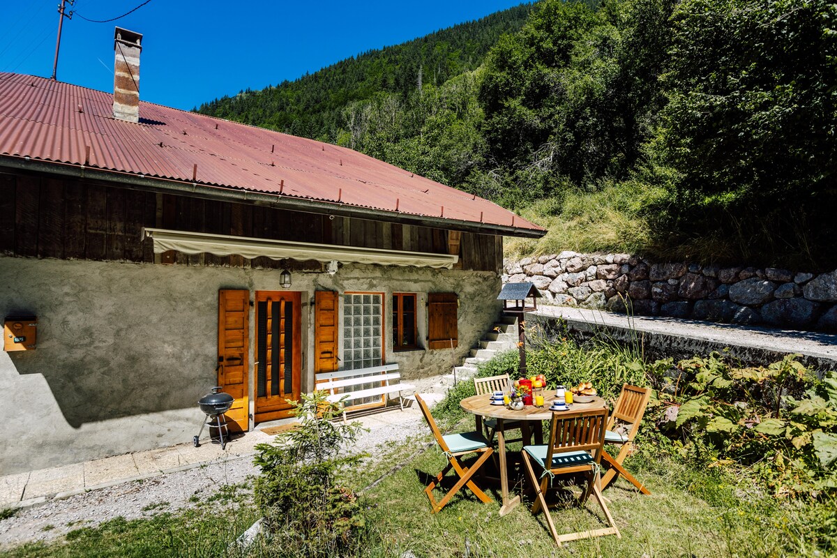 An outdoor dining area is set with a round table and several wooden chairs. Brightly colored dishes and drinks adorn the table. The rustic exterior of the barn features wood shutters and a charming entrance, framed by greenery and mountain views under a clear blue sky.
