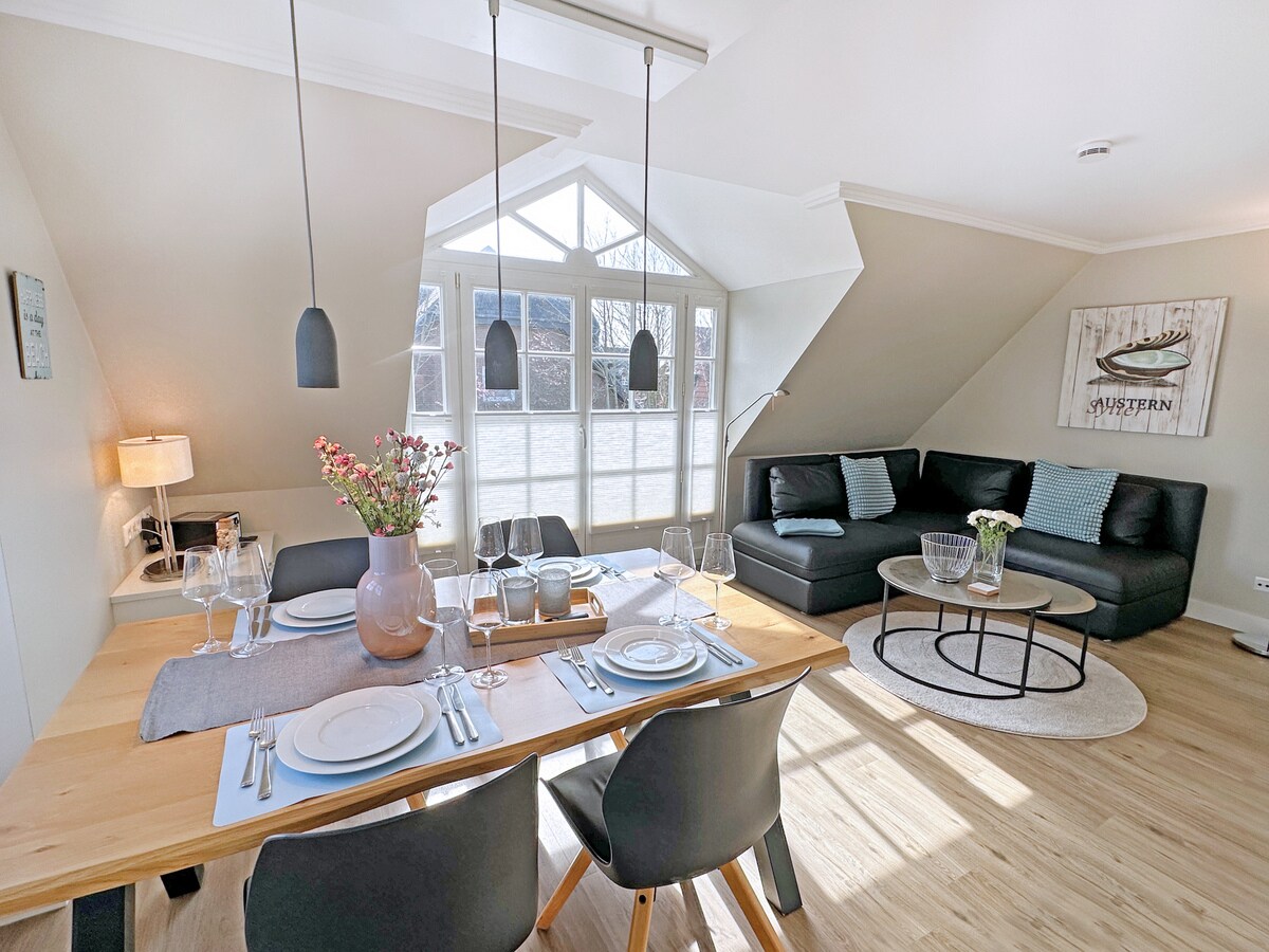 A modern living area features a bright dining table set for meals, accompanied by black chairs. A comfortable sofa provides seating. Large windows allow natural light to fill the space, enhancing the airy feel. Decorative elements include a vase of flowers and wall art.