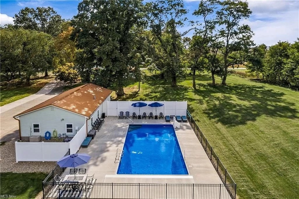 An aerial view of a spacious saltwater pool surrounded by a concrete patio and lounge chairs. Blue umbrellas provide shade, while the property is framed by lush green lawns and trees. A building housing amenities is visible nearby.