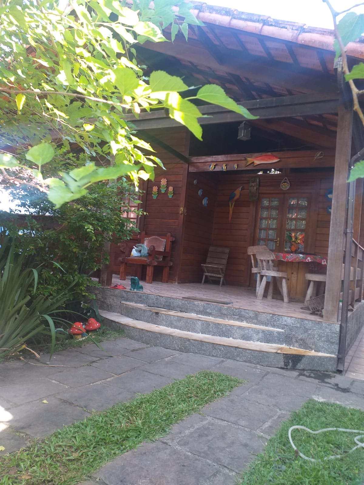 A charming wooden house entrance is framed by vibrant greenery. There are stone steps leading up to a covered porch, which features wooden seating and decorative elements. The surrounding garden includes well-maintained grass and lively plants, creating a welcoming outdoor space.