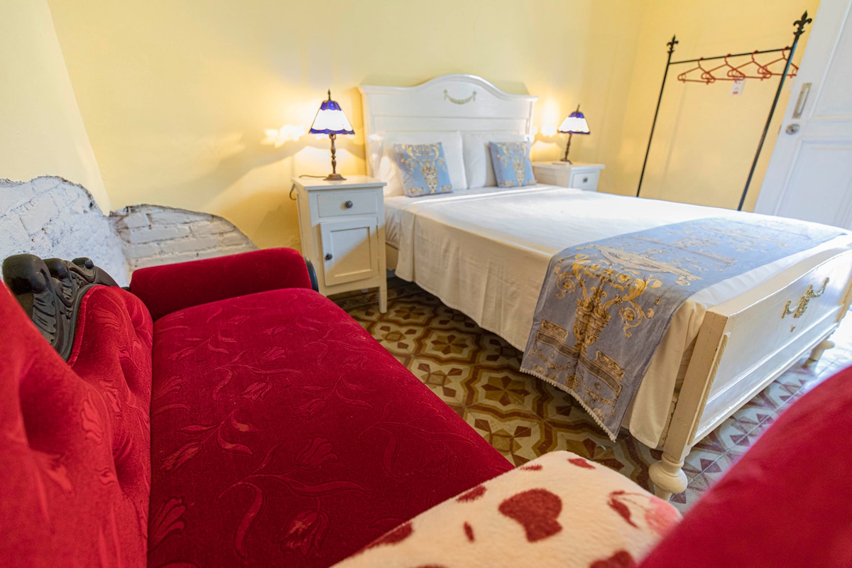 A cozy bedroom features a spacious bed dressed in crisp white linens and accented with a blue decorative cover. A red couch sits nearby, paired with two traditional bedside tables, each holding a lamp. The patterned floor adds a unique touch to the warm yellow walls.