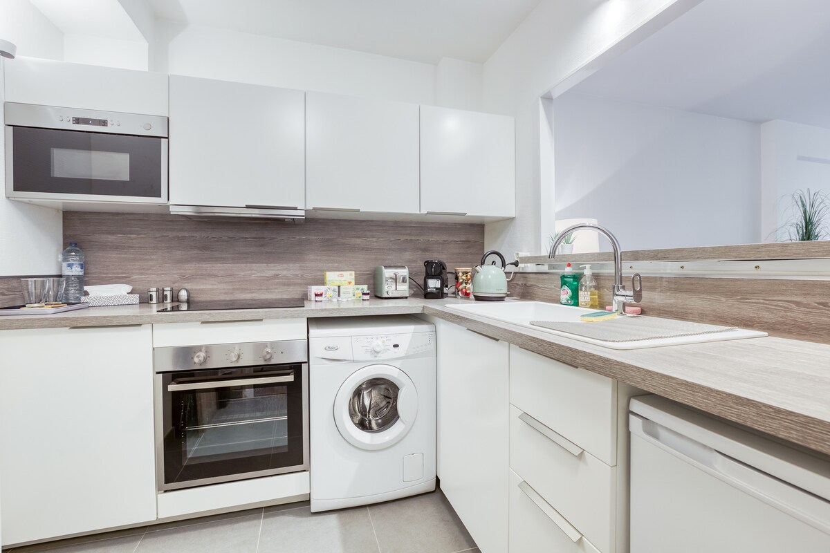 The kitchen is equipped with modern appliances, including an oven, microwave, and washing machine. A neutral color palette of white cabinetry complements the wooden countertop. Kitchenware and utensils are organized neatly, providing a functional cooking space.