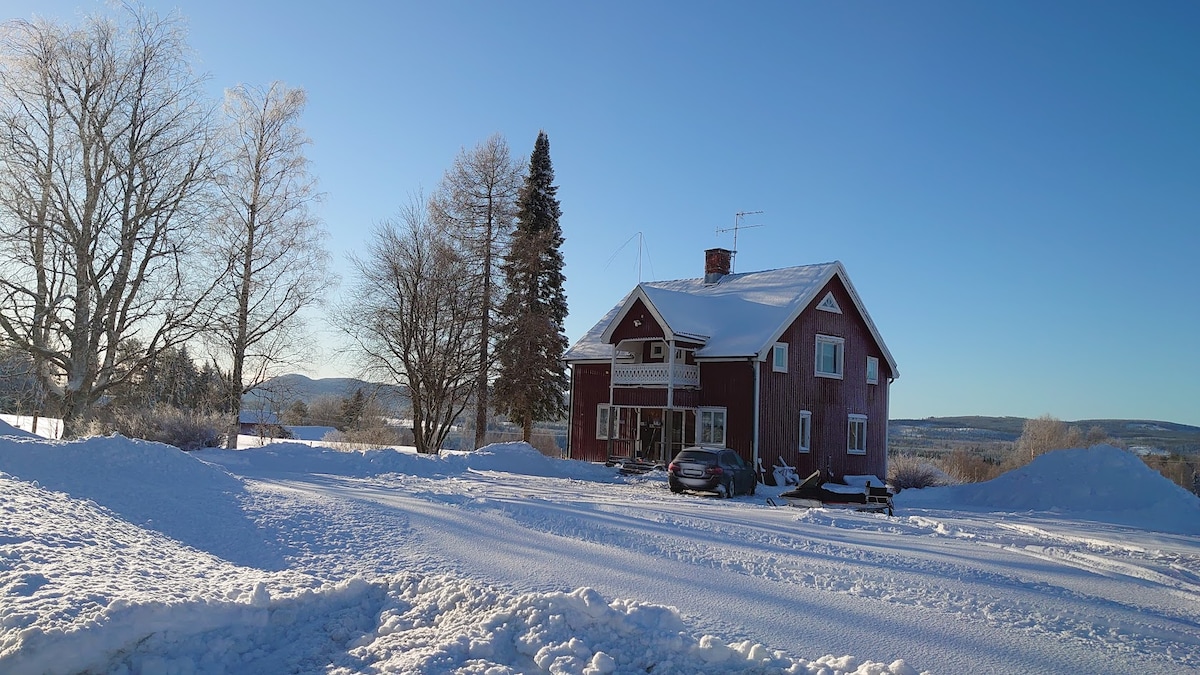 A traditional two-story red house is surrounded by snow-covered grounds, with evergreen trees framing the property. The clear blue sky creates a bright atmosphere, and a car is parked on the spacious driveway, providing easy access to the entrance.