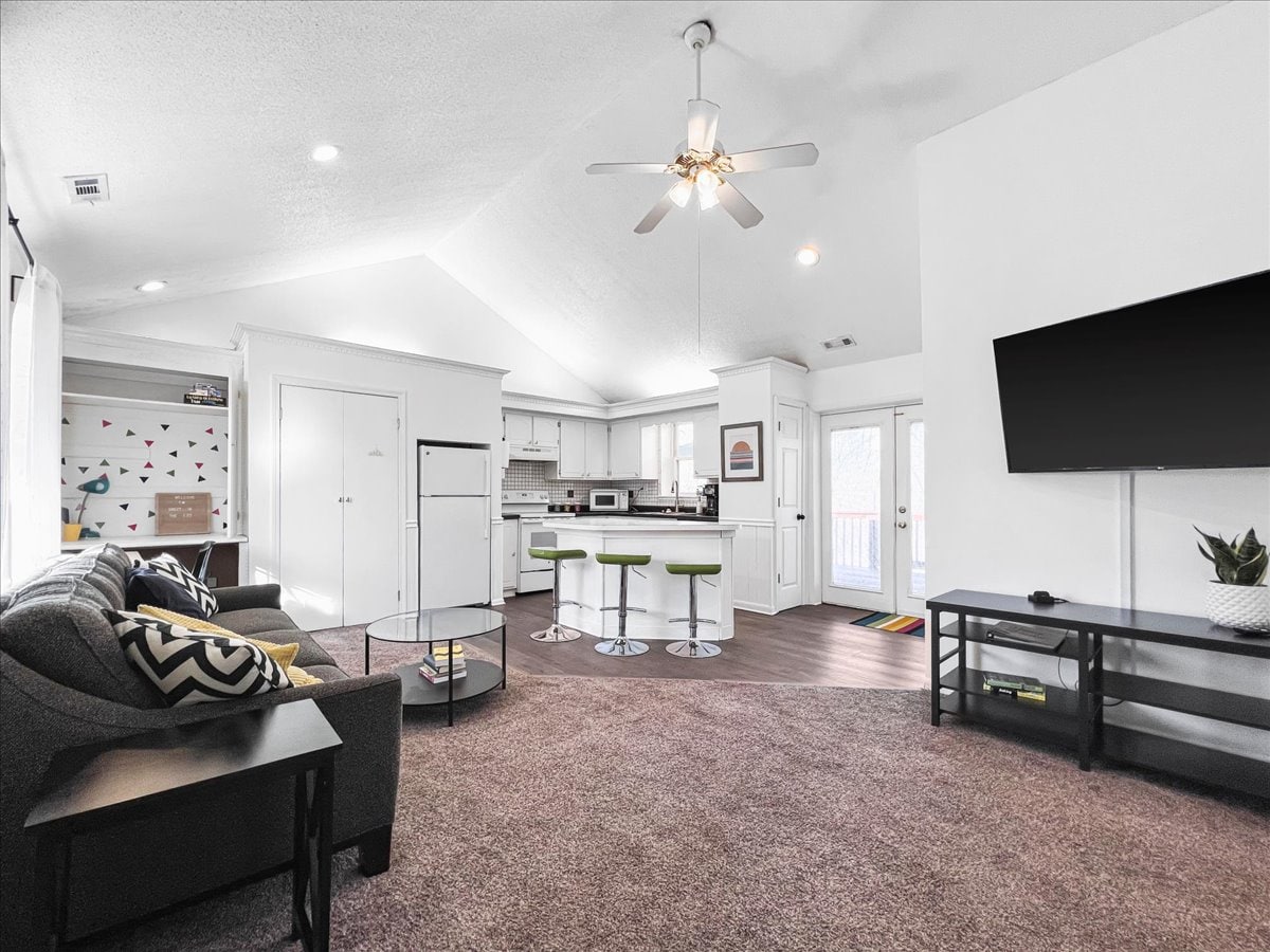 The living area features a plush couch and a modern coffee table, with a cabinetry unit visible in the background. A ceiling fan provides airflow. The kitchen showcases a breakfast bar with seating, and large sliding doors lead to an outdoor space.
