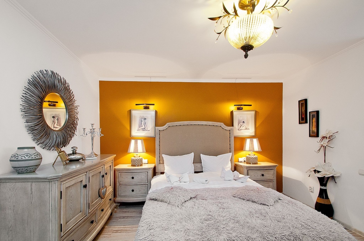 A spacious bedroom is highlighted by an orange accent wall and elegant lighting. A king-size bed, adorned with soft white linens and a textured blanket, is flanked by two nightstands with lamps. Decorative mirrors and framed art pieces enhance the room's charm.