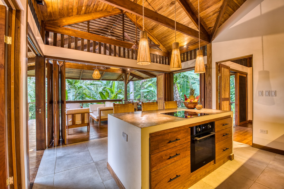 The kitchen features a spacious island with wooden cabinetry and modern appliances, surrounded by wooden beams and pendant lighting. Large glass doors open to a terrace area filled with tropical greenery, enhancing the indoor-outdoor flow of the space.