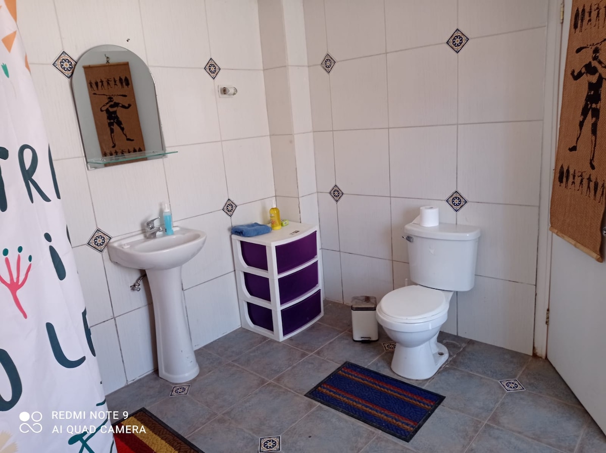 A clean bathroom features white tiled walls and a grey tiled floor. A small mirror is mounted above a sink beside a colorful storage unit with purple drawers. A toilet is positioned next to the sink, with toiletries arranged neatly on the counter.