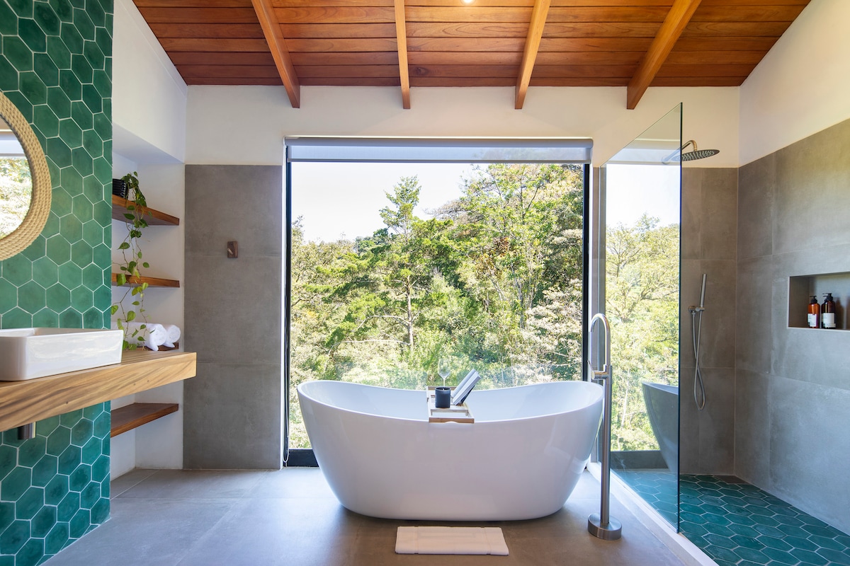 The bathroom features a freestanding tub positioned near a large window, offering views of the surrounding greenery. A modern shower with glass enclosure is visible, along with a circular mirror above a wooden sink. Green hexagonal tiles add a contemporary touch to the space.