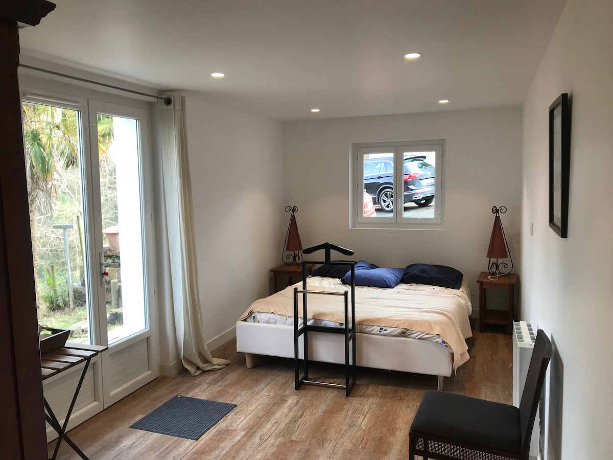 A spacious room features a comfortable bed with neutral bedding and a window allowing natural light to enter. Two bedside tables with lamps are placed beside the bed, and a folding chair is positioned nearby. A view of the exterior is visible through the window.