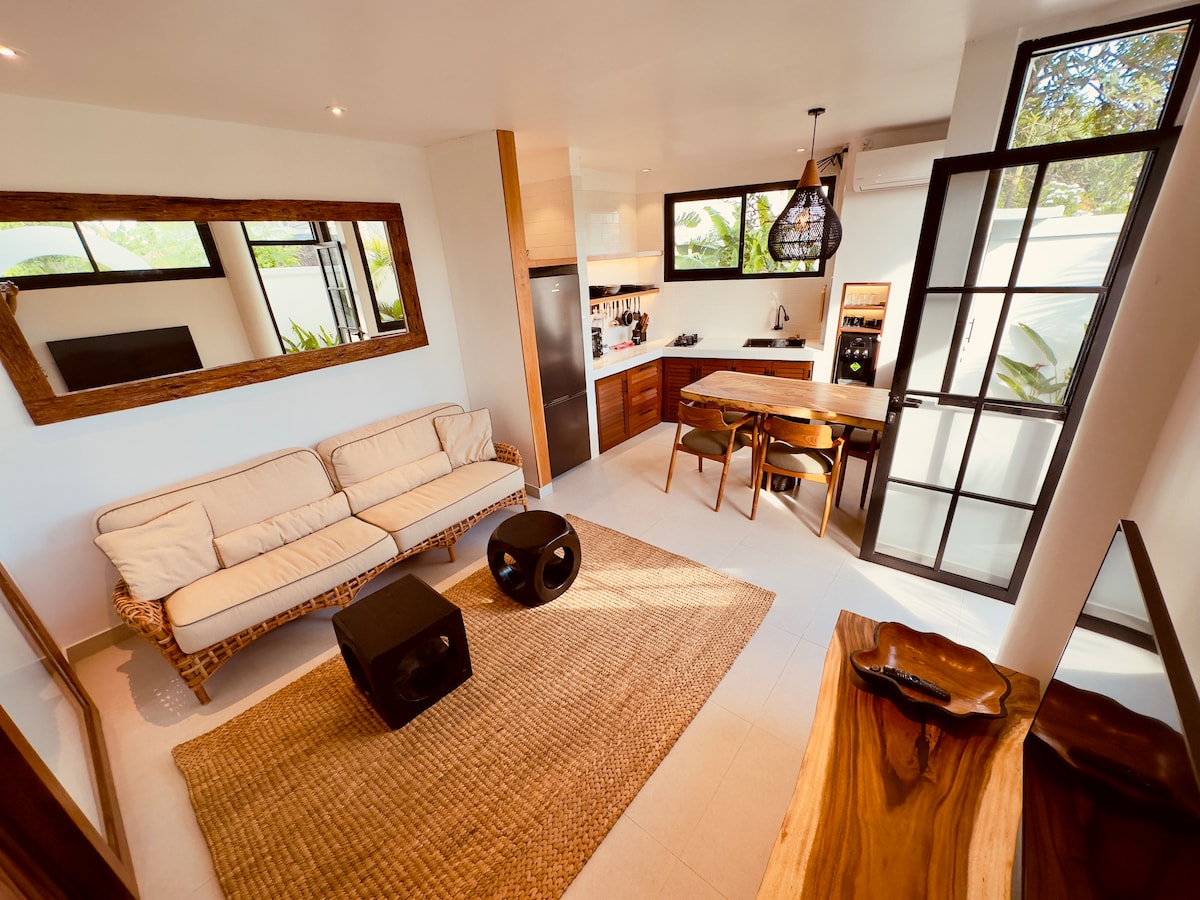 A modern living area features a comfortable light-colored sofa and two dark, round coffee tables. Natural light floods the space through large windows. An open kitchen area is equipped with sleek appliances, while a textured area rug adds warmth to the clean, minimalist design.