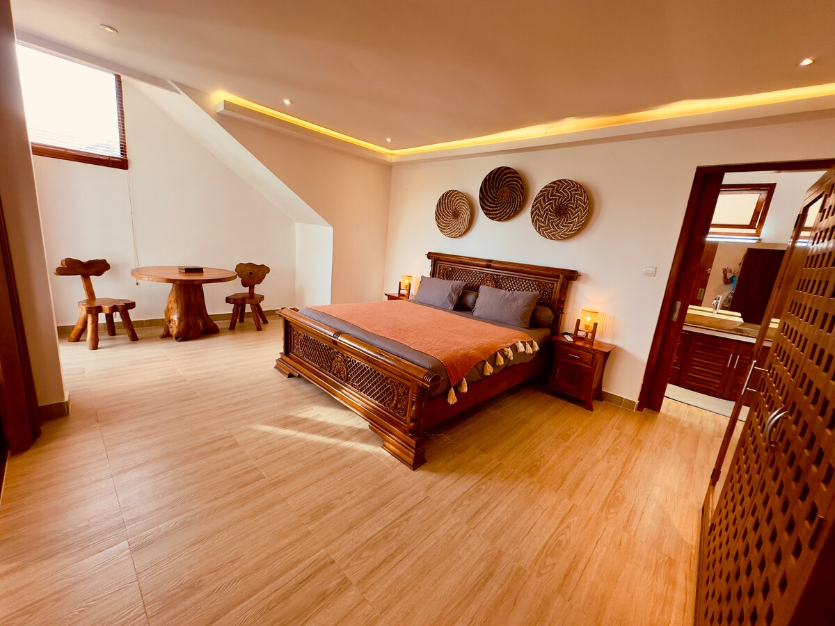 A spacious sleeping area features intricate wooden furnishings, including a large bed with decorative pillows. A round wooden table and two unique chairs are positioned nearby. Soft lighting accents the room, creating a calm and welcoming atmosphere.