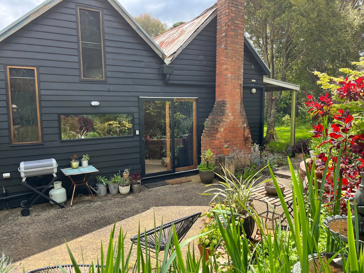 A charming exterior of a cottage is showcased, featuring a dark timber facade and a prominent brick chimney. Lush greenery surrounds the space, with potted plants lining the entrance. A barbecue grill is positioned on the patio, inviting outdoor cooking.