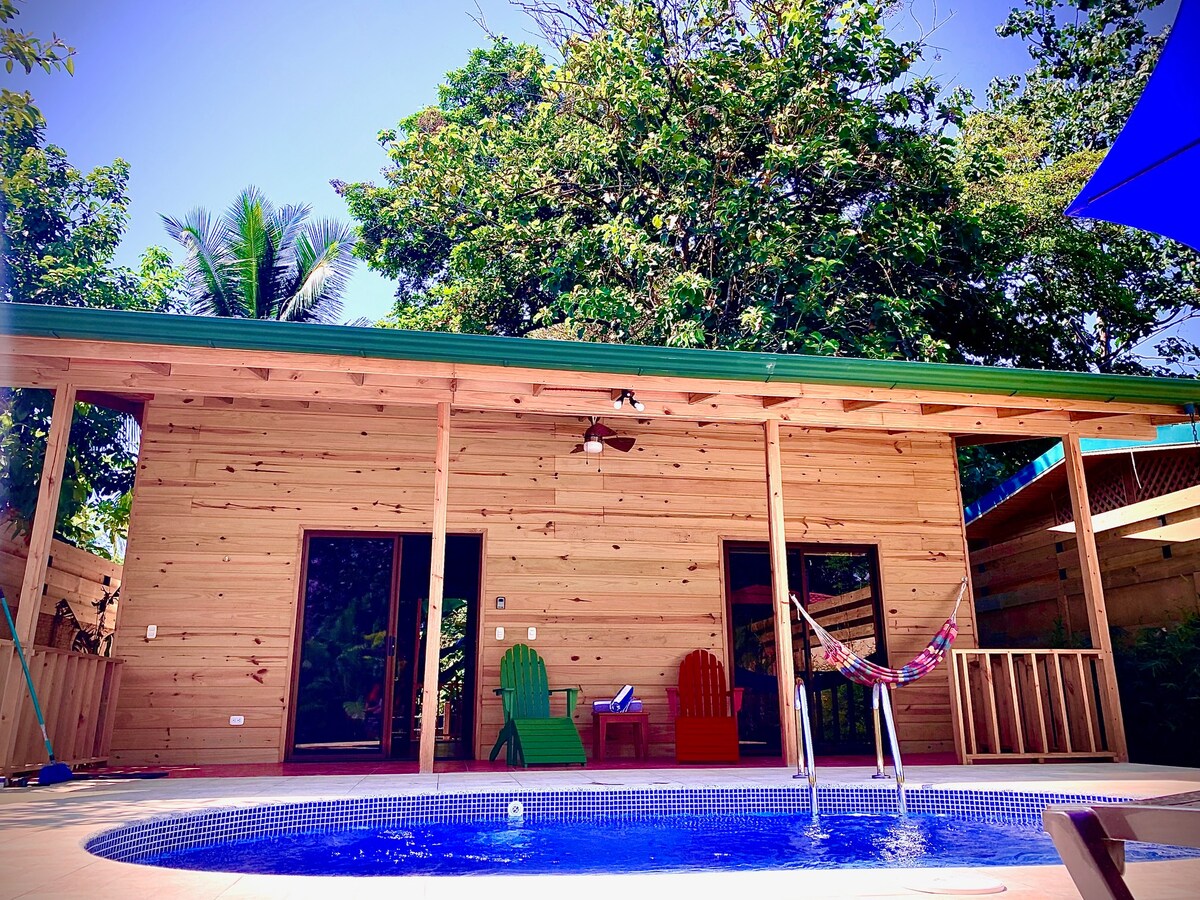 A wooden structure with a green roof is presented alongside a circular pool. Two red and green chairs are positioned nearby, with a hammock hanging in the background. Lush trees provide shade, enhancing the tropical ambiance of the outdoor space.