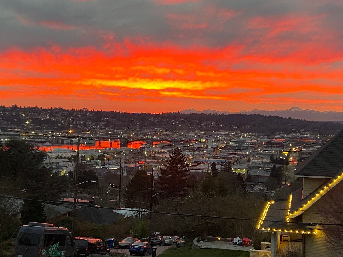 A vibrant sunset blankets the sky in shades of red and orange, casting reflections over the city below. Dark silhouettes of buildings and distant mountains create a striking contrast against the colorful horizon.