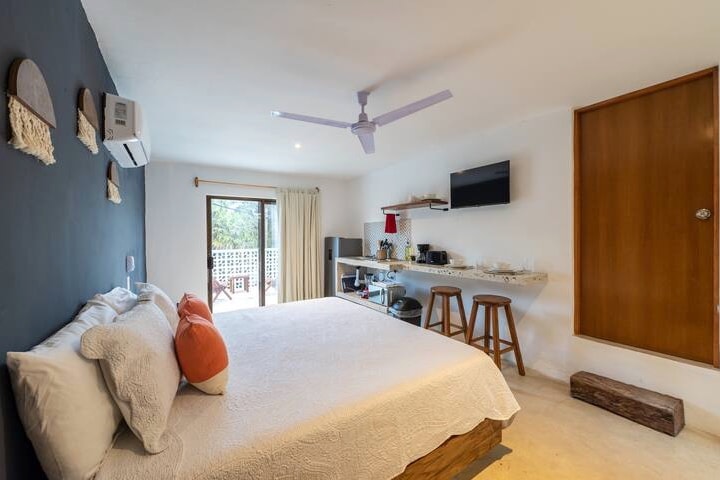 A bright and airy studio features a spacious bed, accented with decorative pillows. A ceiling fan enhances airflow. A wooden shelf displays kitchen amenities, while a Smart TV is mounted on the wall. Large glass doors open to a private terrace, inviting natural light.