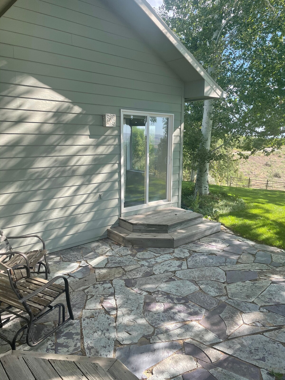 An outdoor seating area features two metal chairs positioned on a stone patio, leading to a glass sliding door. Sunlight filters through nearby trees, casting dappled shadows on the patio's natural stone surface, which complements the home's light-colored siding.
