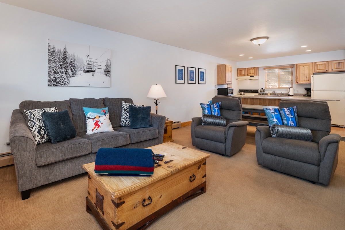 A comfortable living area features two cushioned chairs and a spacious sofa adorned with decorative pillows. A wooden coffee table occupies the center. Bright kitchen cabinetry can be seen in the background, enhancing the open layout of the space.