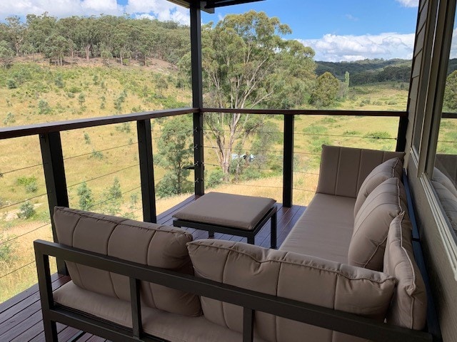A covered balcony features a comfortable seating area with plush, light-colored cushions. A small table is placed in the center, and expansive views of the surrounding natural landscape can be seen beyond the railing, showcasing trees and grassy fields.