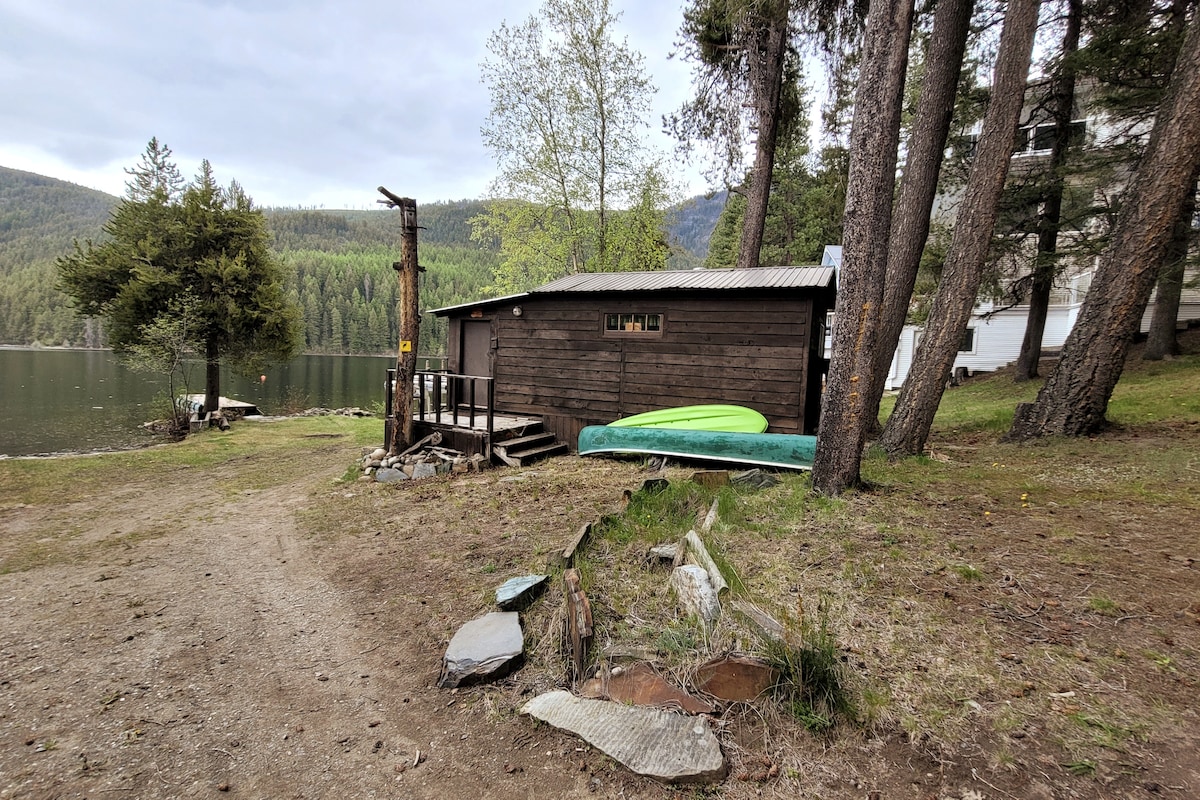 A cozy cabin is nestled along the water's edge, surrounded by tall trees. A grassy area leads to a private deck with a gentle slope toward the lake. Two kayaks are stored against the cabin, inviting outdoor activities. The serene landscape is ideal for relaxation.
