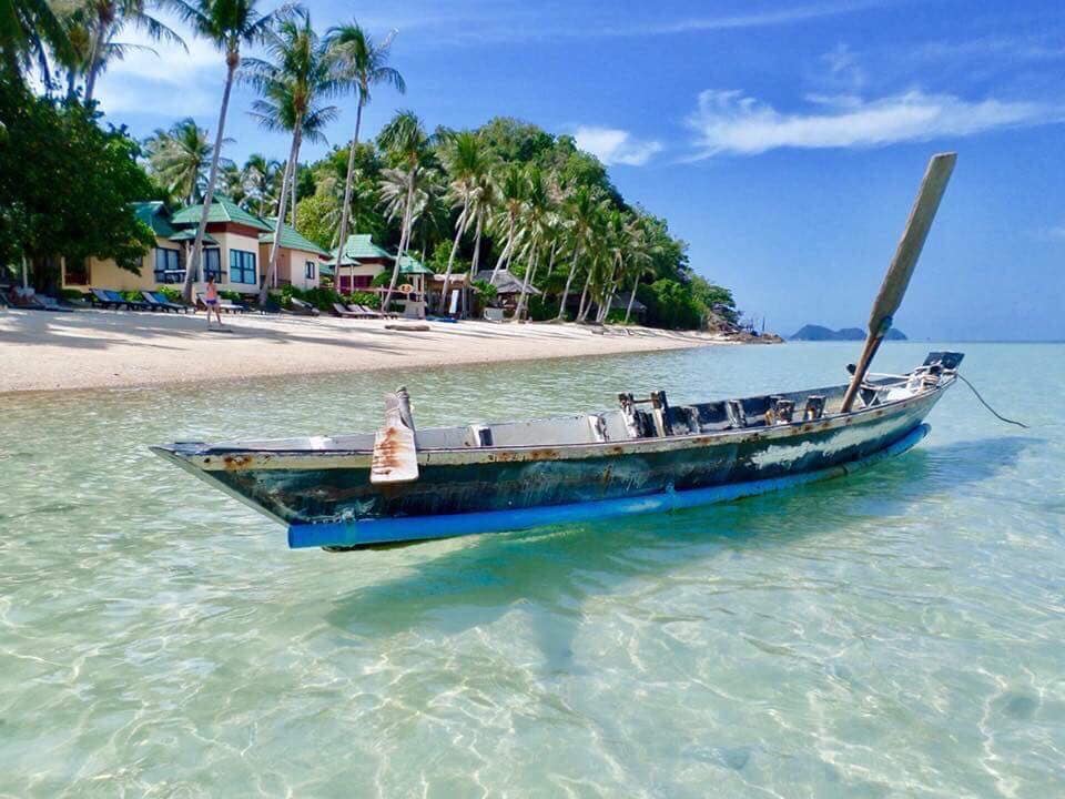 An abandoned boat rests in clear turquoise waters, gently bobbing near a sandy beach. Palm trees line the shore, with a bungalow visible in the background, surrounded by lush greenery under a clear blue sky.