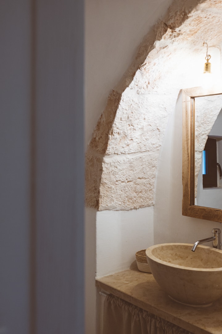 A rustic bathroom features an arched stone wall, illuminated by warm light. A round, natural stone sink sits on a simple countertop, complemented by a wooden-framed mirror above. Soft fabric drapes beneath the countertop, adding a touch of warmth to the space.