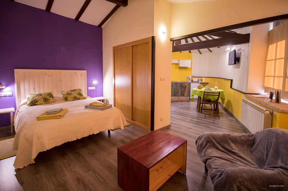 A spacious studio apartment features a cozy sleeping area with a light-colored bedspread, complemented by a vibrant purple accent wall. A small wooden table is positioned nearby, while the kitchen-dining area is visible in the background, showcasing modern amenities and natural lighting.