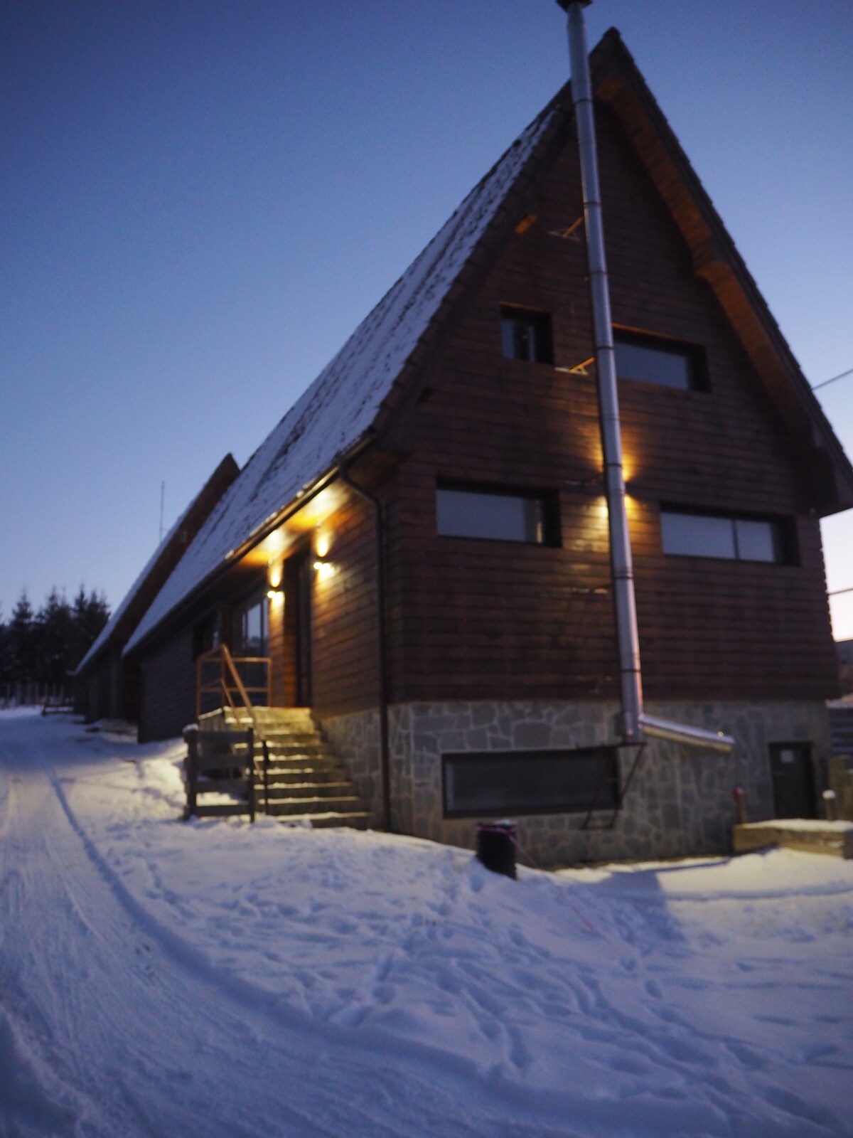 A wooden chalet is set against a twilight sky, showcasing a steeply pitched roof and large windows. Soft lighting outlines the structure, illuminating steps leading to the entrance. Snow blankets the ground, creating a serene winter atmosphere around the property.