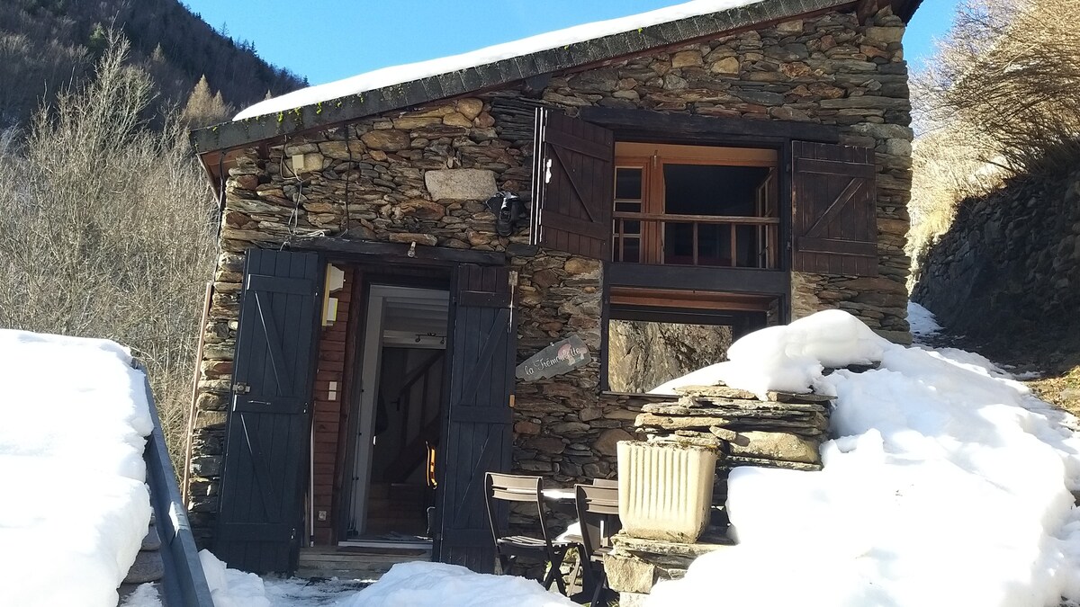 A rustic stone cottage is nestled in a snowy landscape, showcasing wooden shutters and a sloped roof. A small seating area is visible in front, surrounded by scattered snow and framed by trees and mountains in the background.