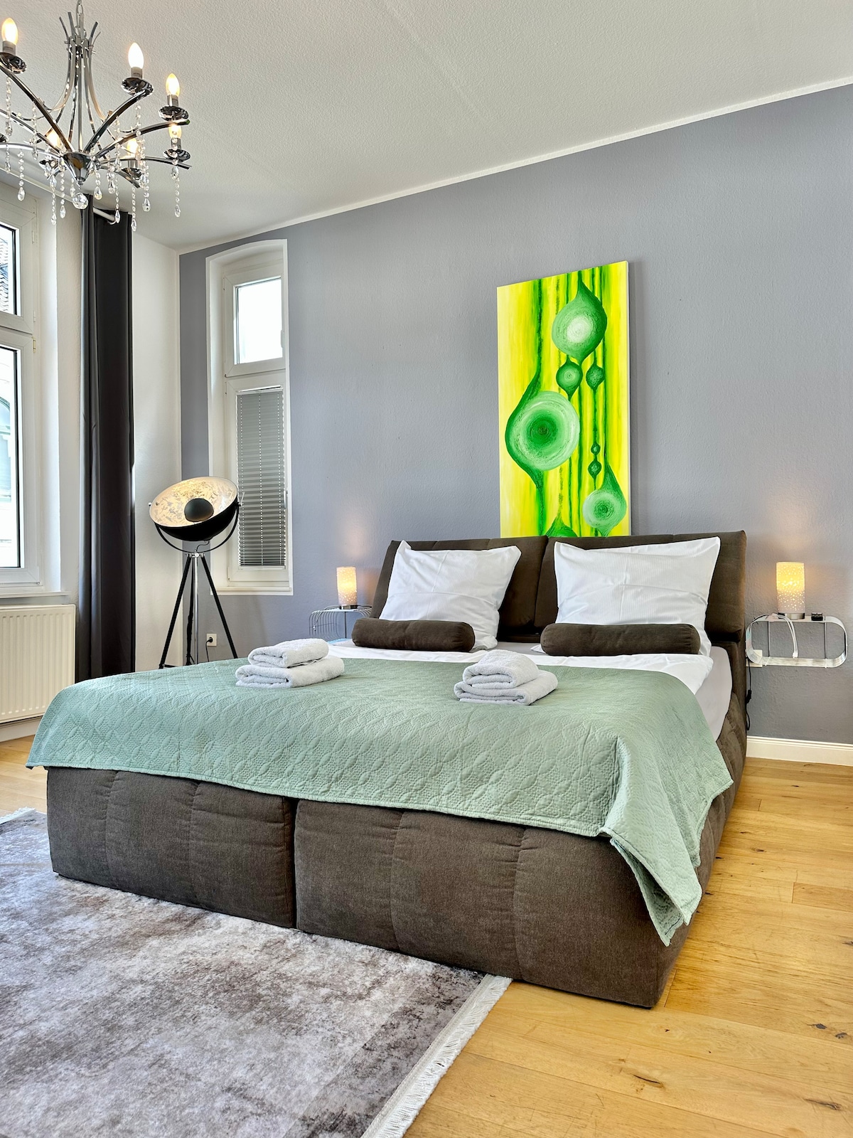 A spacious bedroom featuring a large bed with a soft green throw. Vibrant green artwork hangs above the bed, complemented by bedside lamps. Natural light filters through large windows, enhancing the warm wooden flooring and stylish chandelier overhead.