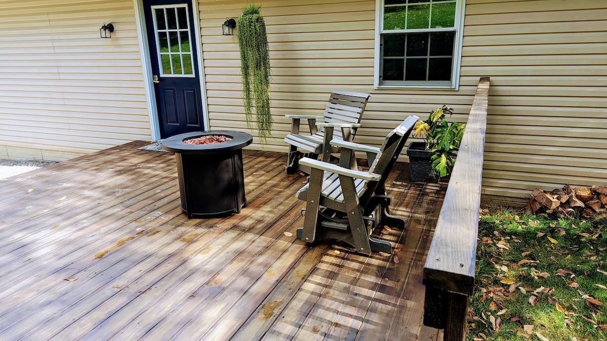 A wooden deck features two Adirondack chairs positioned around a circular fire pit, creating a cozy outdoor space. Lush greenery is present in a hanging planter alongside the deck, while a door with a black frame leads into the home.