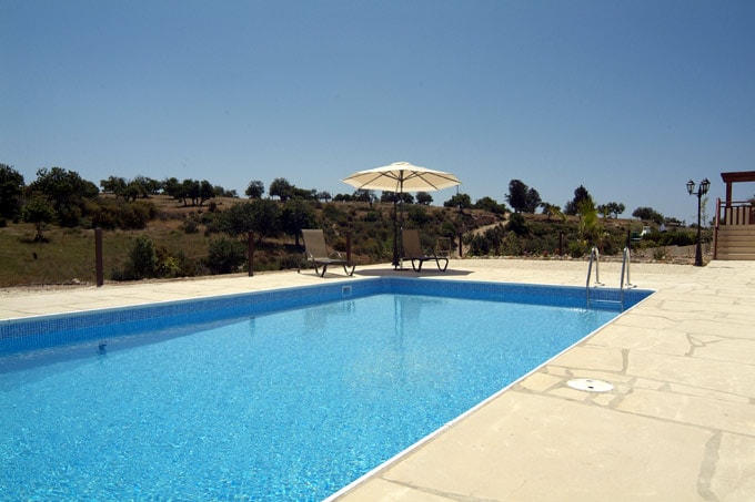 A clear blue swimming pool is set within a sun-drenched patio area. Two sun loungers are positioned under a large umbrella, providing shade. Expansive views of open countryside are visible in the background, contributing to a serene outdoor environment.