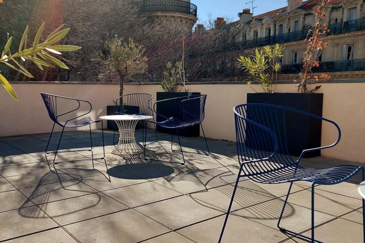 A terrace is depicted with four blue wire chairs surrounding a small table. Potted olive trees and other greenery are arranged in planters, creating an inviting outdoor space. Sunlight filters gently over the tiled surface, casting soft shadows.
