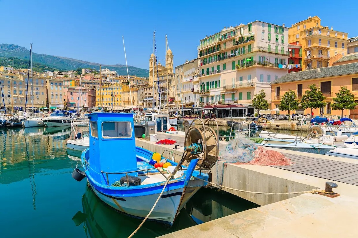 A vibrant blue fishing boat is docked at a marina, surrounded by numerous boats reflecting in calm waters. Historic buildings with varied colors line the waterfront, and distant mountains provide a scenic backdrop under a clear blue sky.
