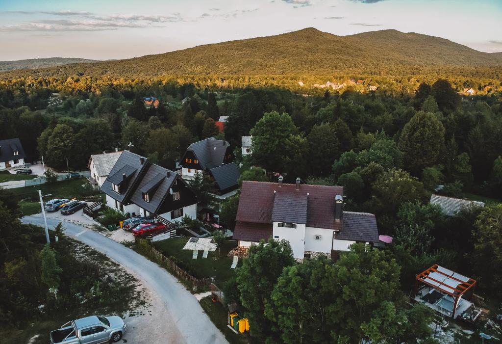 An aerial view captures a serene neighborhood nestled amidst lush greenery and rolling hills. The landscape features houses with varying roof styles, surrounded by trees, with a winding road and parked cars hinting at a tranquil environment close to nature.