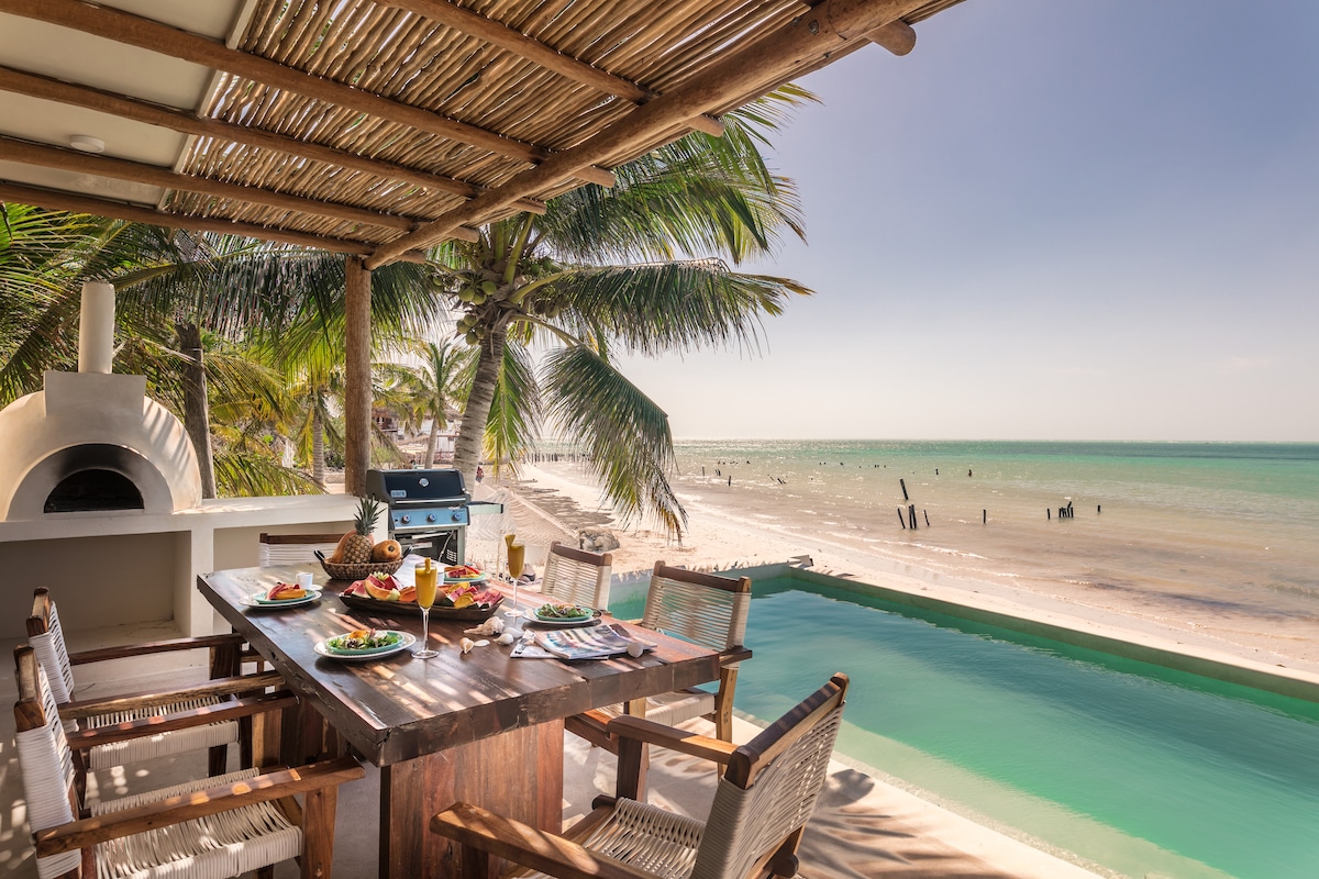 An outdoor dining area with a large wooden table is set with colorful dishes and drinks. A pool edges the space, while the sandy beach and calm sea extend in the background under a clear sky, accented by swaying palm trees.