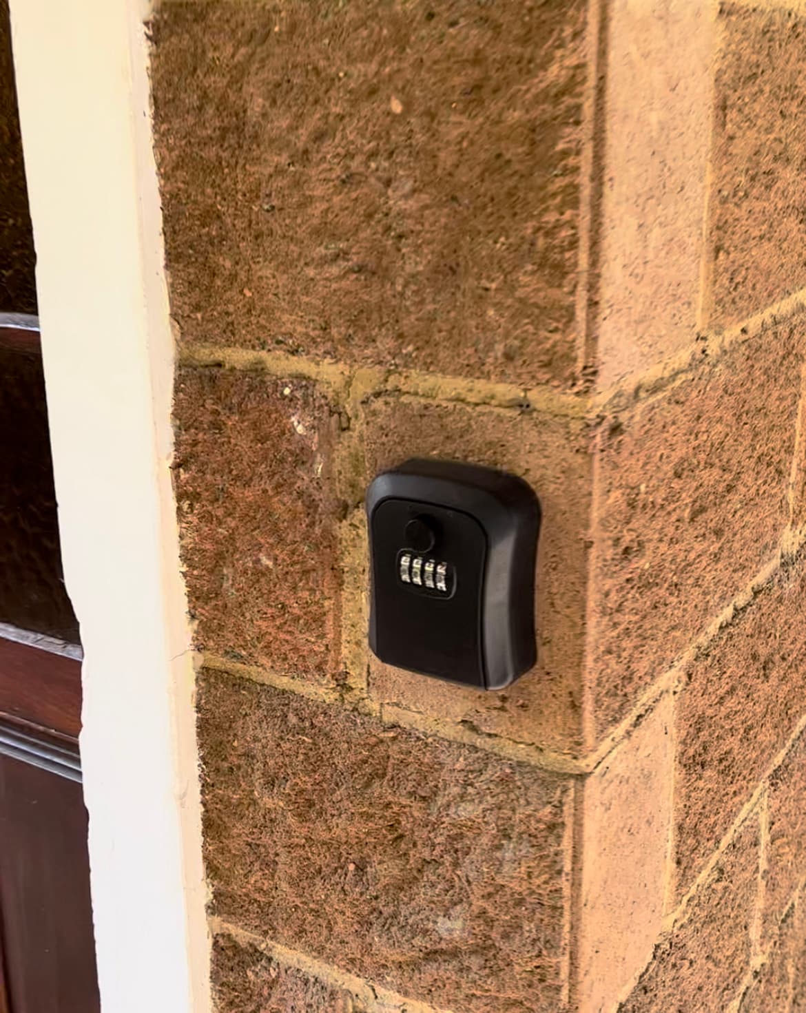 A secure keyless entry system is mounted on a textured stone wall, adjacent to a wooden door. The combination lock features numbered dials, providing easy access for guests. The surrounding area is illuminated by natural light, highlighting the simplicity and functionality of the entry design.