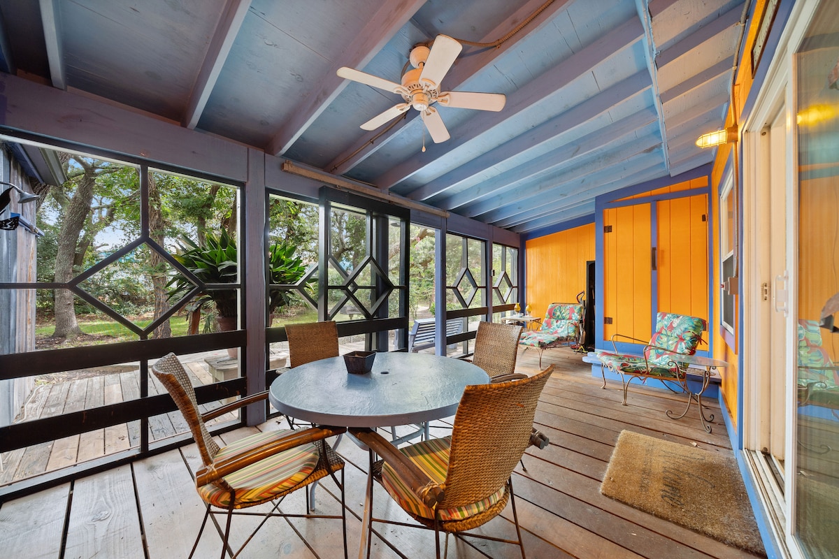 A spacious screened-in porch features a round table surrounded by four chairs, creating an ideal space for relaxation. A ceiling fan is mounted overhead, and colorful seating arrangements are visible at the side. Natural light filters in through the surrounding screens, offering views of the outdoors.