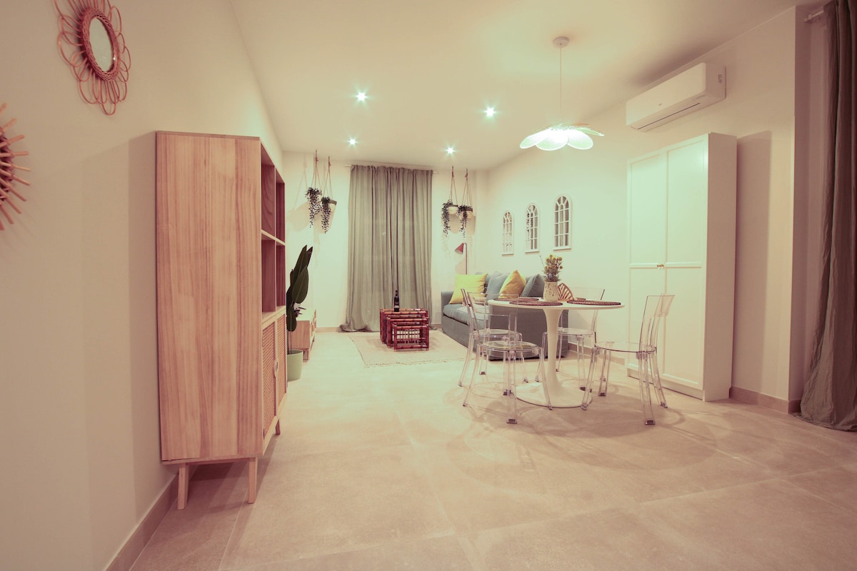 A spacious living area is shown with soft lighting. A comfortable grey sofa is positioned against a wall adorned with decorative windows. A glass dining table surrounded by transparent chairs is visible. Plants hang from the ceiling, contributing to a relaxed ambiance.