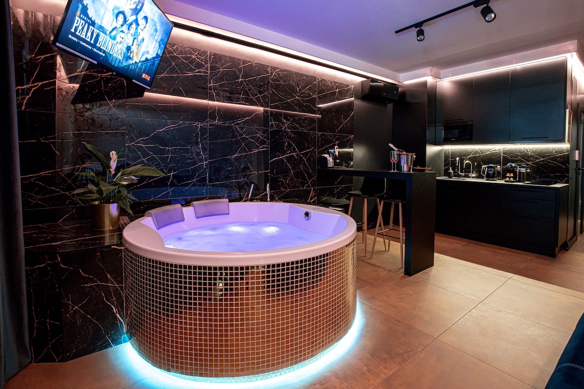 A modern jacuzzi is surrounded by elegant dark marble walls, featuring subtle lighting that enhances the space. A flat-screen TV is mounted above, and a well-equipped kitchen area is positioned nearby, complete with sleek cabinetry and minimalist bar stools.