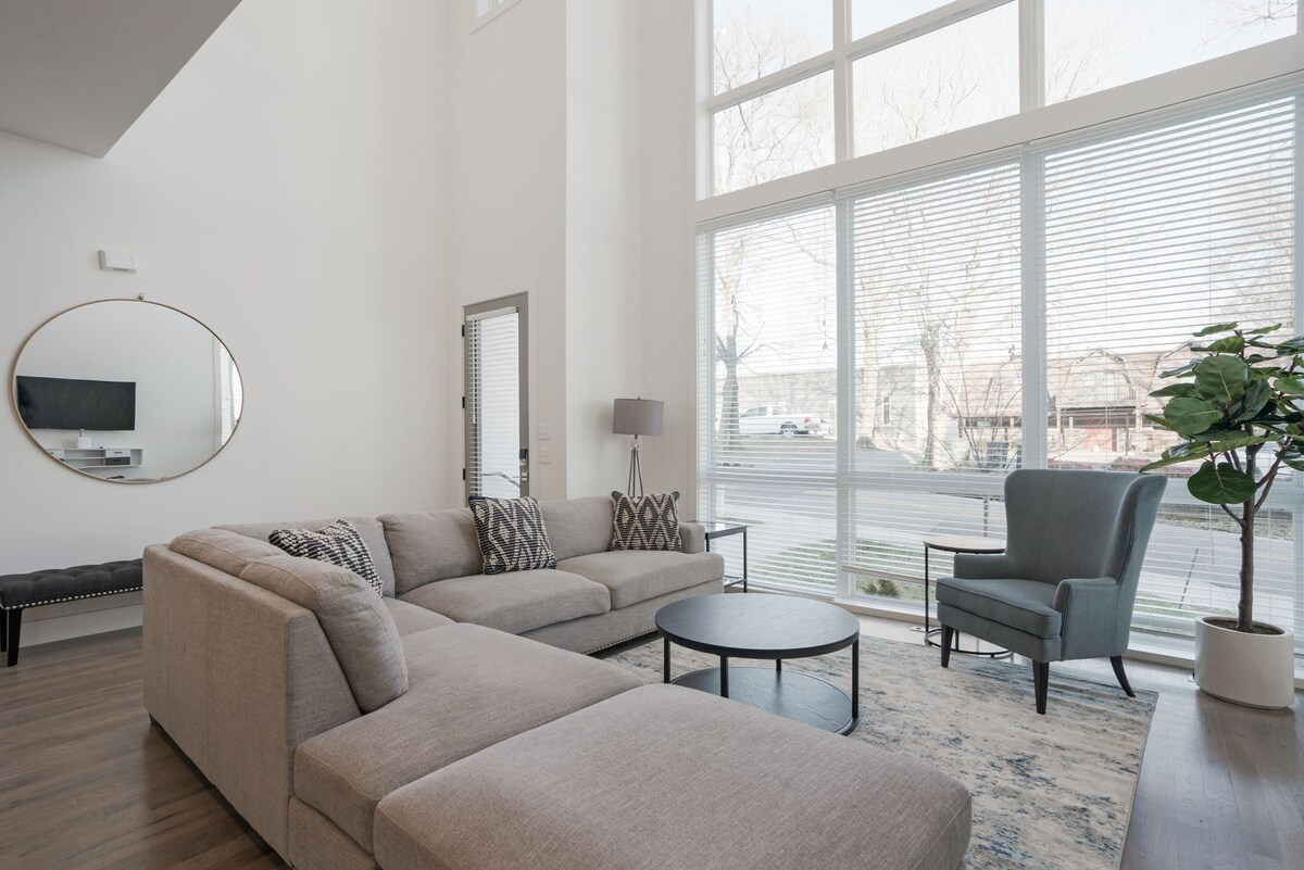 A spacious living area features large windows allowing ample natural light. A comfortable sectional sofa is complemented by an accent chair, and a round coffee table sits in the center. A flat-screen TV is mounted on the wall, while a decorative mirror enhances the room's openness.