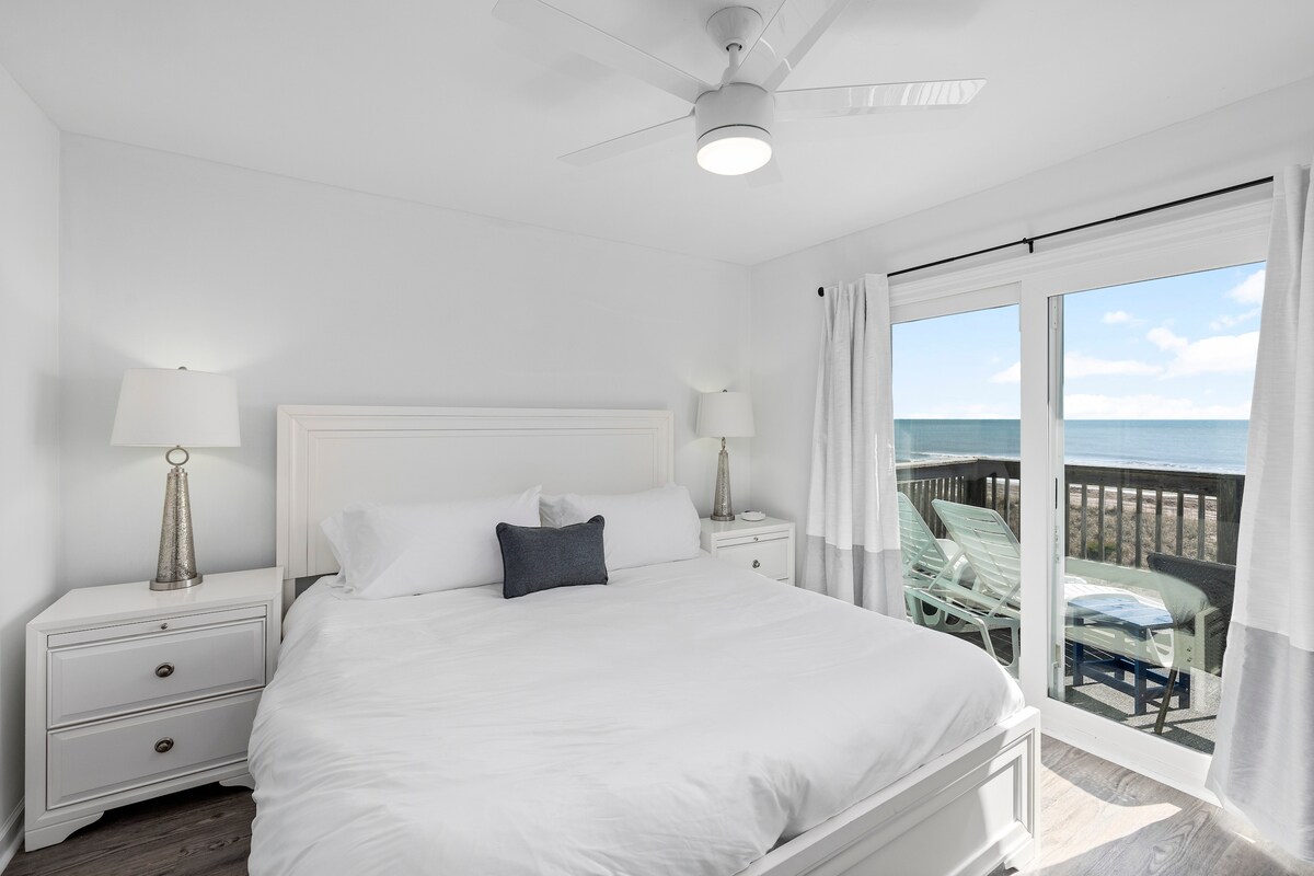 A bright and airy master bedroom is shown with a spacious bed dressed in white linens. Two bedside lamps illuminate the nightstands, and large windows offer views of the ocean. The sliding glass door opens onto a private deck featuring seating for enjoying the scenery.