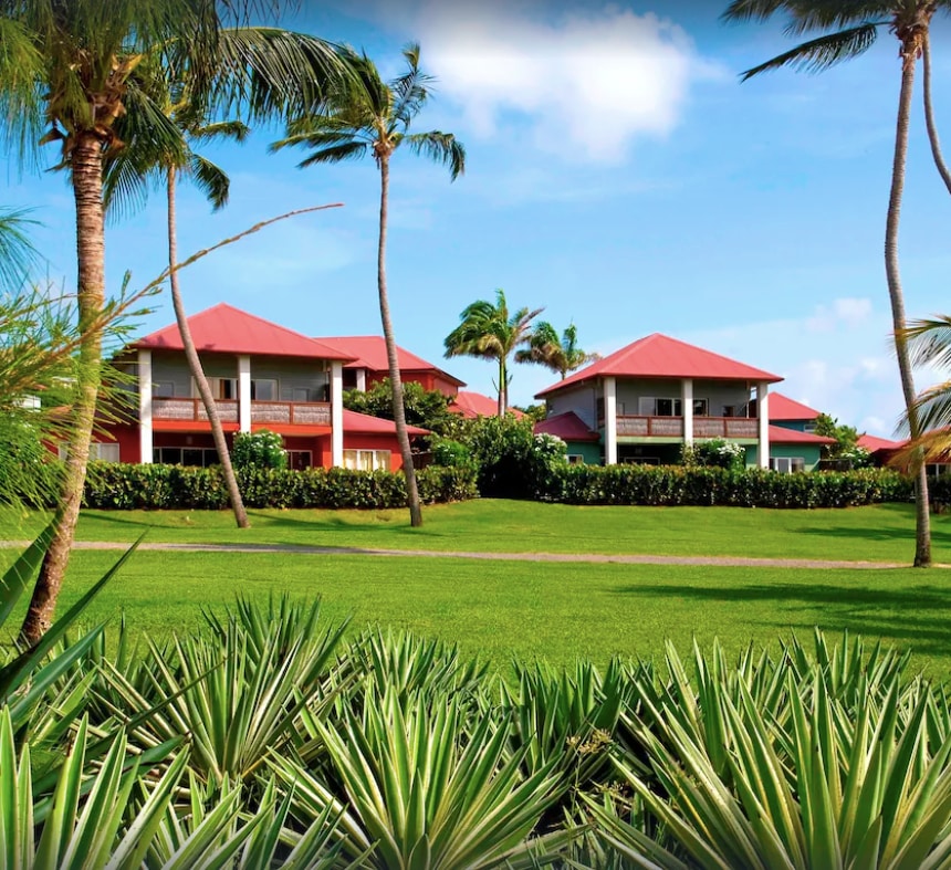 The image features well-maintained grounds with lush greenery in the foreground and palm trees swaying gently. Three spacious villas with red roofs and white pillars are visible in the background, set against a bright blue sky, offering a serene tropical ambiance.