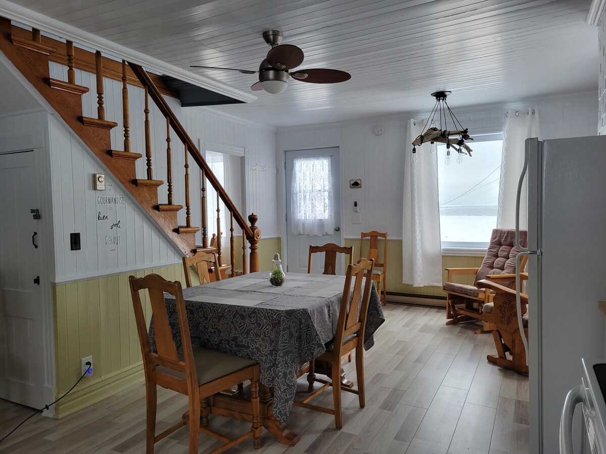 A bright and open living area features a wooden dining table surrounded by chairs, with natural light entering through large windows. A cozy rocking chair is positioned nearby, while the wooden staircase adds charm to the space. Light-colored walls enhance the airy feel.