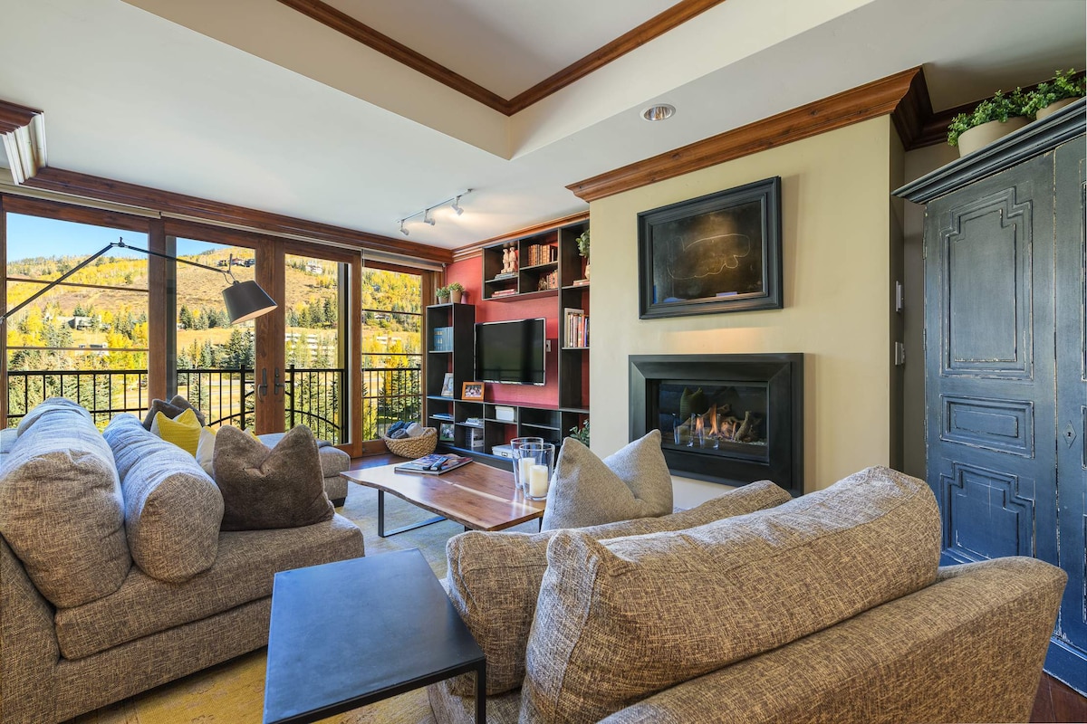 The living area is furnished with plush sofas, providing a comfortable setting for relaxation. A modern television is mounted on the adjacent wall, and a cozy fireplace is featured, framed with decorative elements. Large windows offer scenic views of the surrounding mountains.