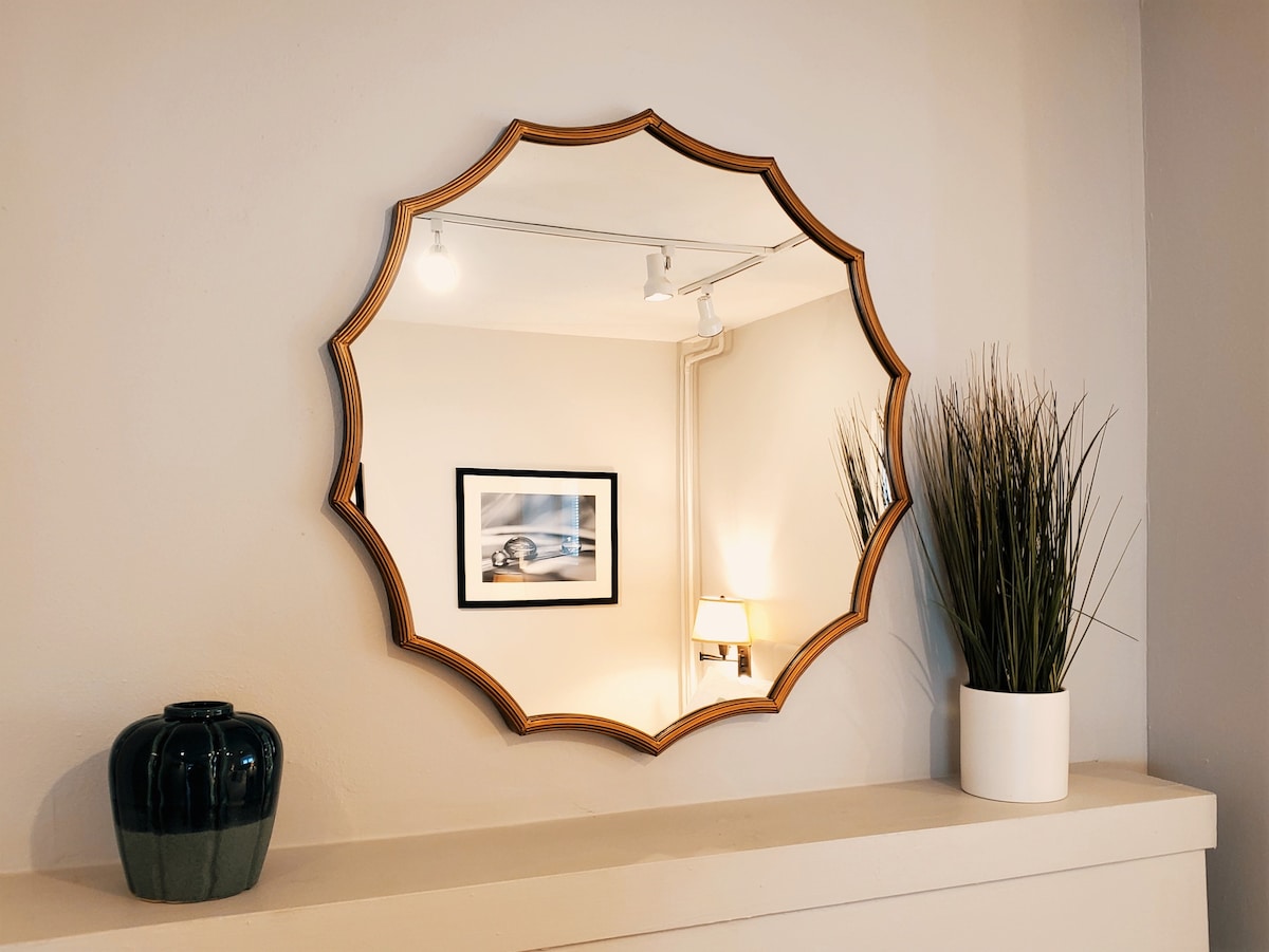 A round mirror with a scalloped wooden frame reflects the room's decor. A black decorative vase sits on the ledge beside a potted plant. Soft lighting illuminates the space, highlighting a framed photograph in the background.
